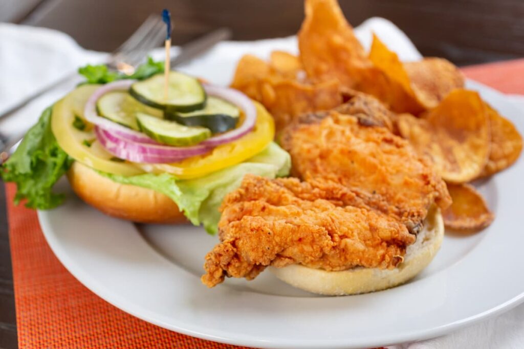 Chick-fil-a serves breakfast in a white plate having burger with crispy chicken cucumber slices, onion slices, capsicum slices, lettuce along with crispy potato chips.