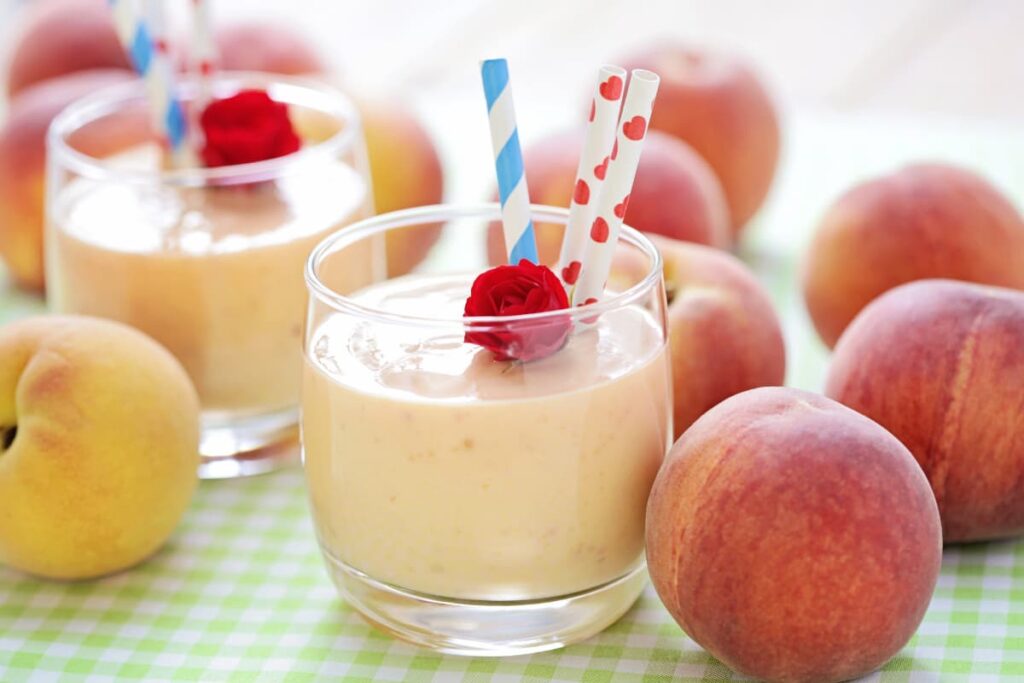 Chick Fil A Peach Milkshake are served in 2 glasses, one small red rose for garnishing along with 3 straws. On the floor there are some peaches around the glasses.