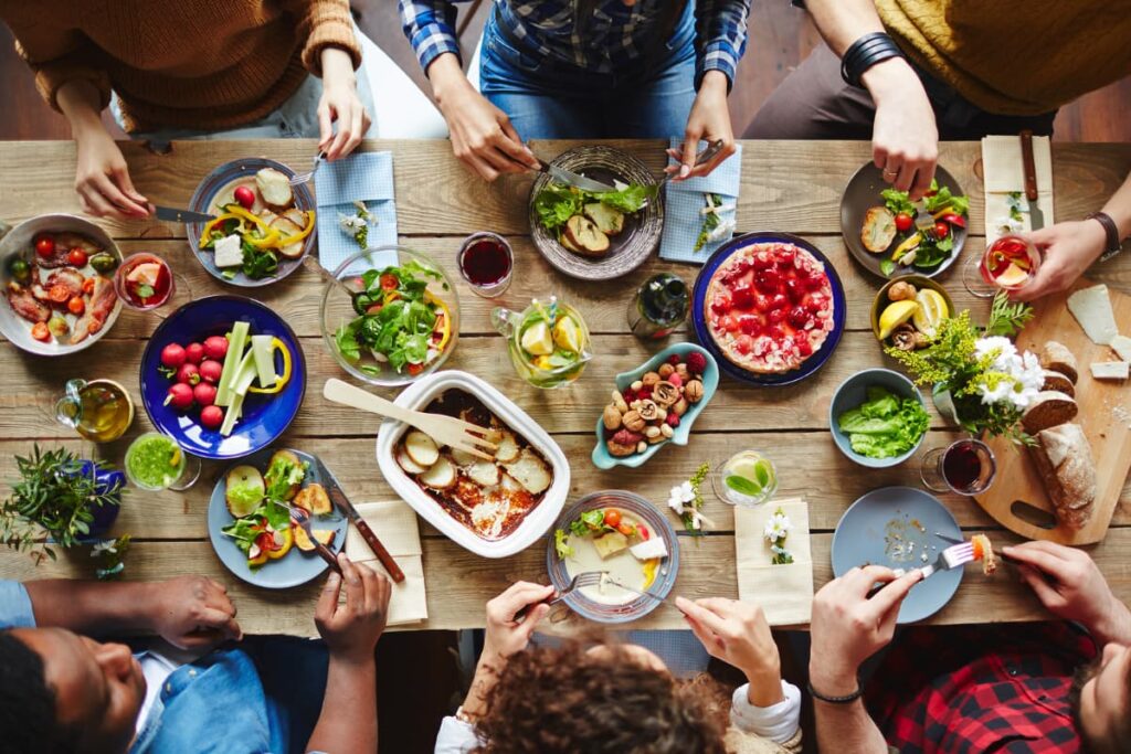 In Chick Fil A Open Dining Room 6 members are seated and started eating. In a brown color table there are lot of food like salads, fruits, boiled vegetables, brown bread, drinks, plates, cups, knives, spoons, forks etc.