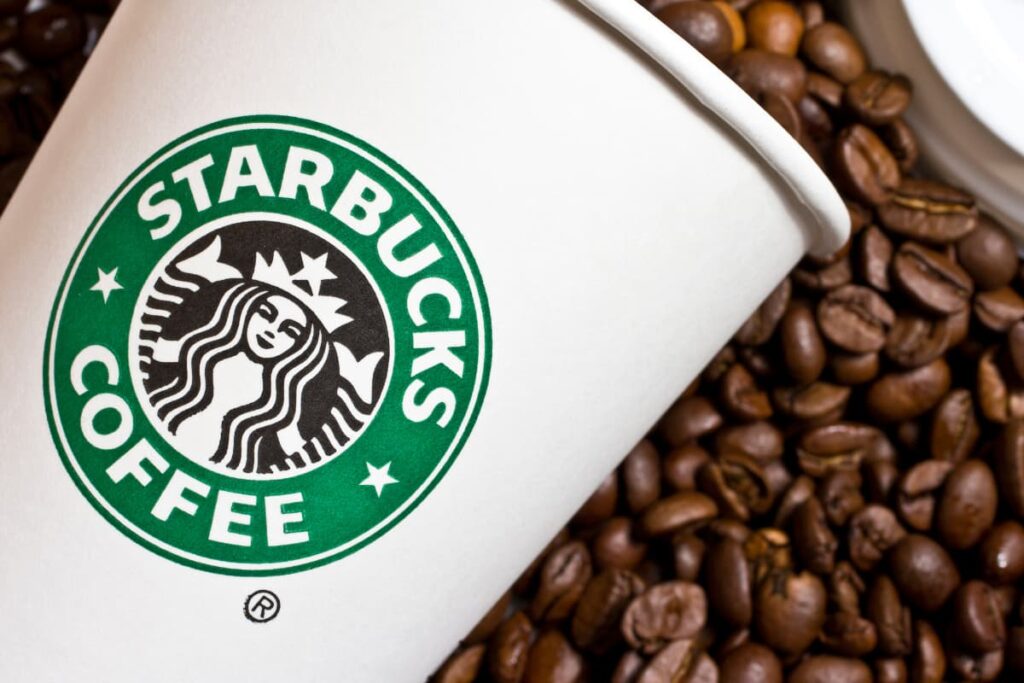 The Starbucks Logo is in a cup and it is green and white color combination. In Starbucks Logo there is a mermaid around it starbucks coffee. Beside the starbucks coffee cup, there are some coffee beans on the floor.