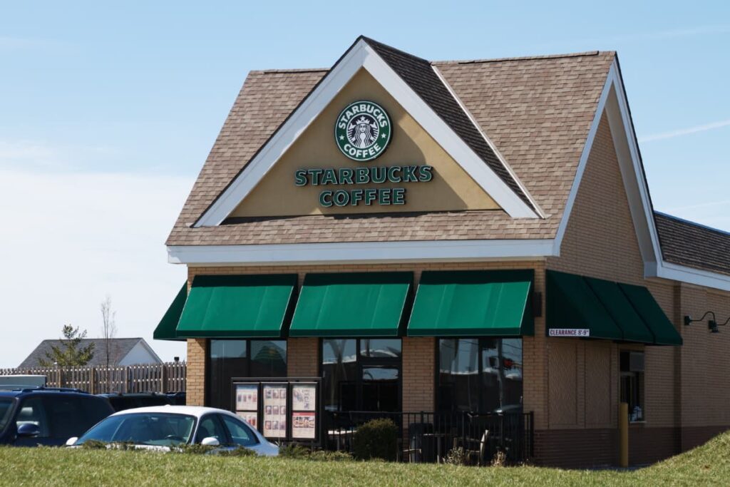 Starbucks owner is Howard Schultz who built Starbucks. Infront of Starbucks some cars are parked, on the top of the building starbucks logo is there below it is mentioned Starbucks coffee. 