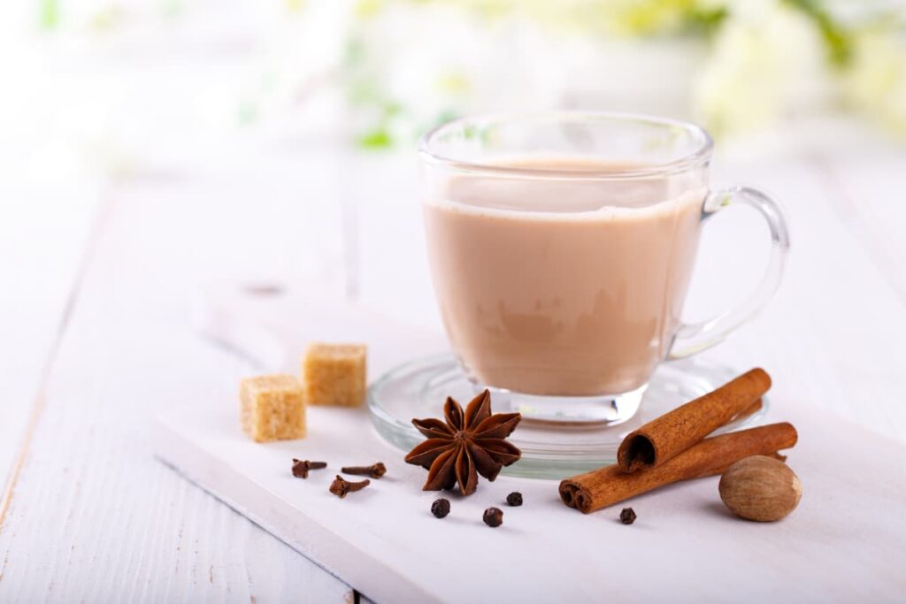 Starbucks Chai Tea serves in a glass cup along with the glass saucer. This starbucks chai tea is kept on a table, beside the chai tea there are 3 cloves, 3 peppers, 2 cinnamon sticks, one anise, 2 sugar cubes and one nut mug.