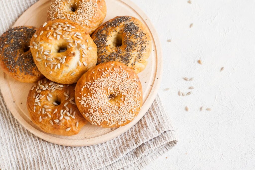 Starbucks Bagels serves in a brown color plate, there are 6 bagels on a plate. All bagels are garnished with some nuts like pumpkin seeds, some sesame seeds etc, and there is a towel under the plate. 