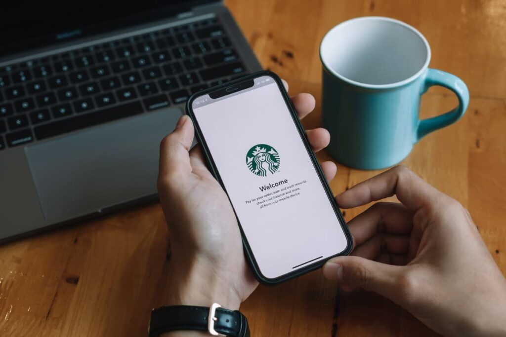 Delete Starbucks Account from your mobile or laptop. On a table there is one laptop, one green color cup, and a phone which is holding a person with his hands and the person wears black watch on his left hand.