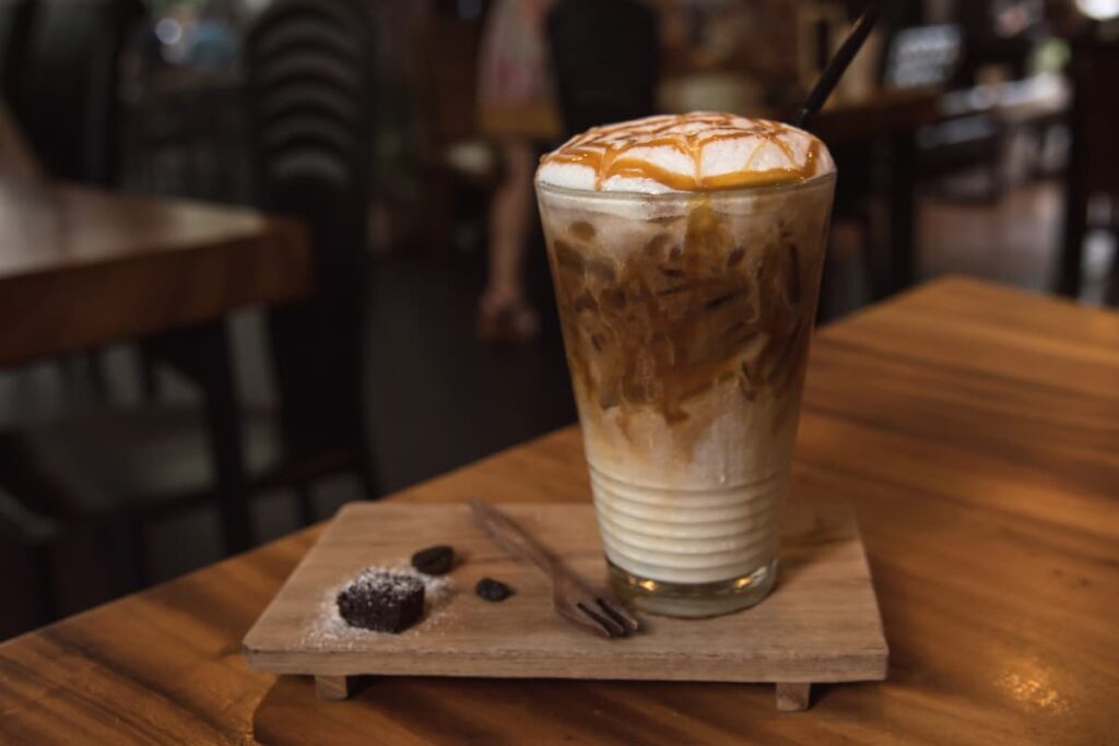 Starbucks Iced Caramel Macchiato can be served in a glass along with the brown color wooden plate, which is also have some pieces of chocolate and it is sprinkled with sugar powder, one fork. Starbucks iced caramel macchiato is garnished with the caramel syrup and all are kept in a brown color table. 