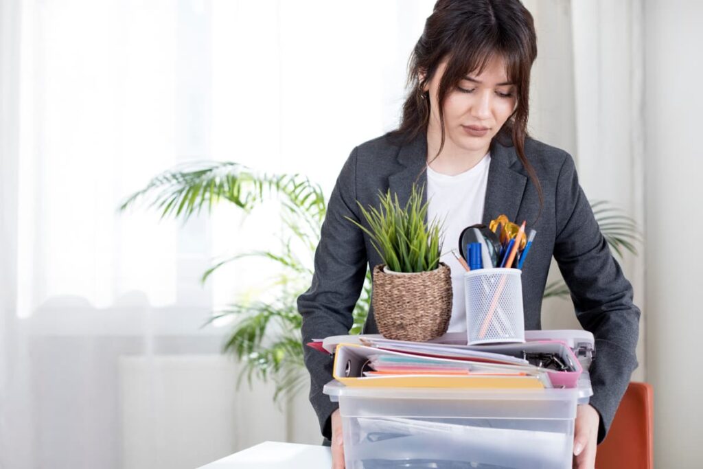 A women who wears ash color coat along with the white t-shirt, holds a box of files and white color metallic pen holder, one small brown color plant on top of it.