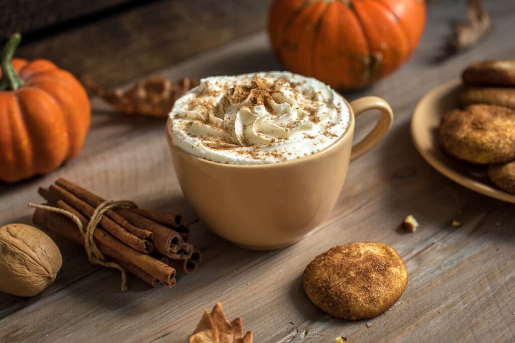 Starbucks Pumpkin Spice is Vegan, because it is used pumpkin and cinnamon. On a table there are 2 orange pumpkins, bunch of cinnamon sticks, one walnut, and a plate of cookies, one cookie is on the table. Pumpkin spice latte is served in a yellow cup, which is garnished with the whipped cream and sprinkled cinnamon powder.