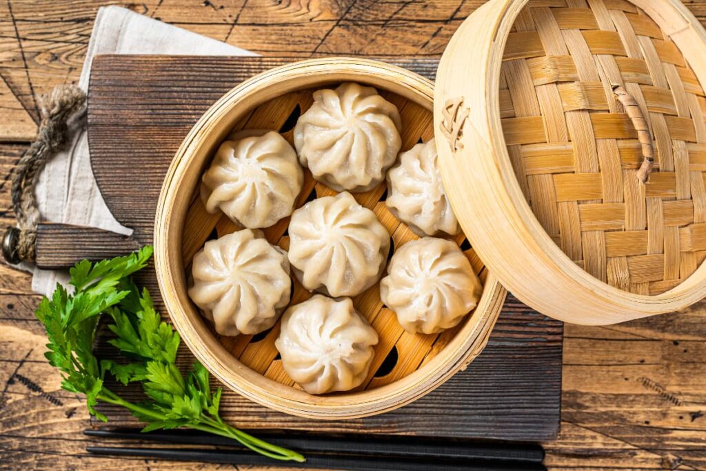 Starbucks Steamers are used to steam momos. Here on a table they serve momos(white in color) in a wooden steamer, besides there are some green leaves.