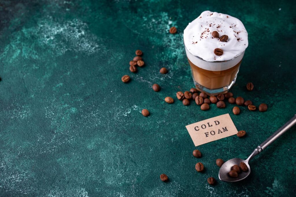 Starbucks Cold Foam can be served in a glass, and it is garnished with coffee beans and coffee powder on top. Starbucks Cold Foam was written on the paper and kept in the floor, beside one spoon of coffee beans, some beans are sprinkled on the floor.