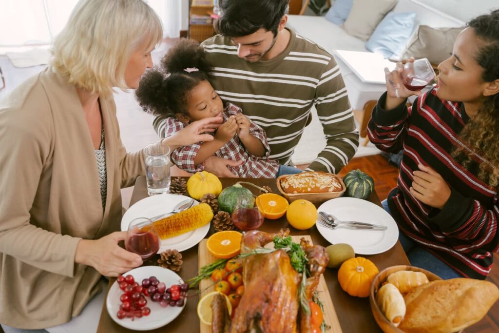 Starbucks Open On Thanksgiving to serve food for families. A family seated around the table, the table includes whole grilled chicken, some red cherries, bread, orange slices, 3 glasses of wine, tomatoes, cake, kiwi, corn, small pumpkins, water, forks, spoons and 2 plates.