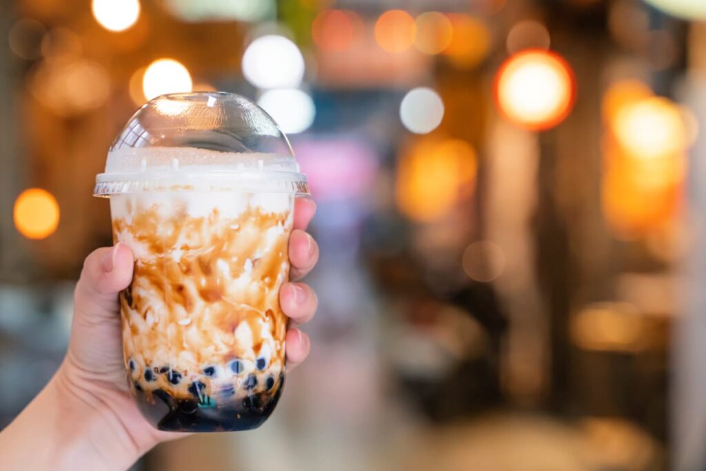 Starbucks Boba can be served in a glass. A person holds starbucks boba at his/her left hand, and the color of the boba is black at the bottom and cream with coffee color at the top.